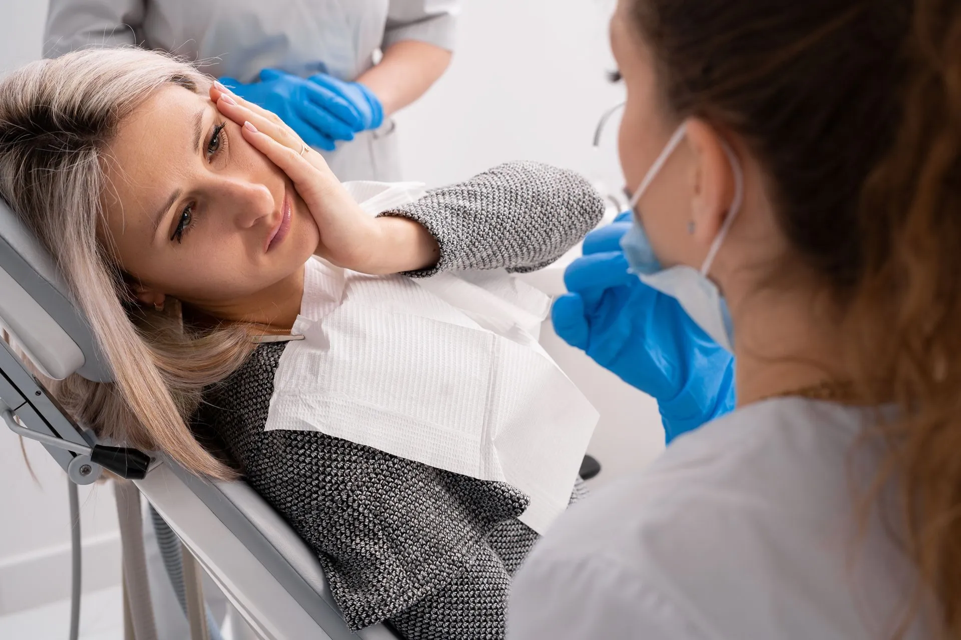 Patient lying in a dental chair holding their jaw while a dentist prepares to assist.