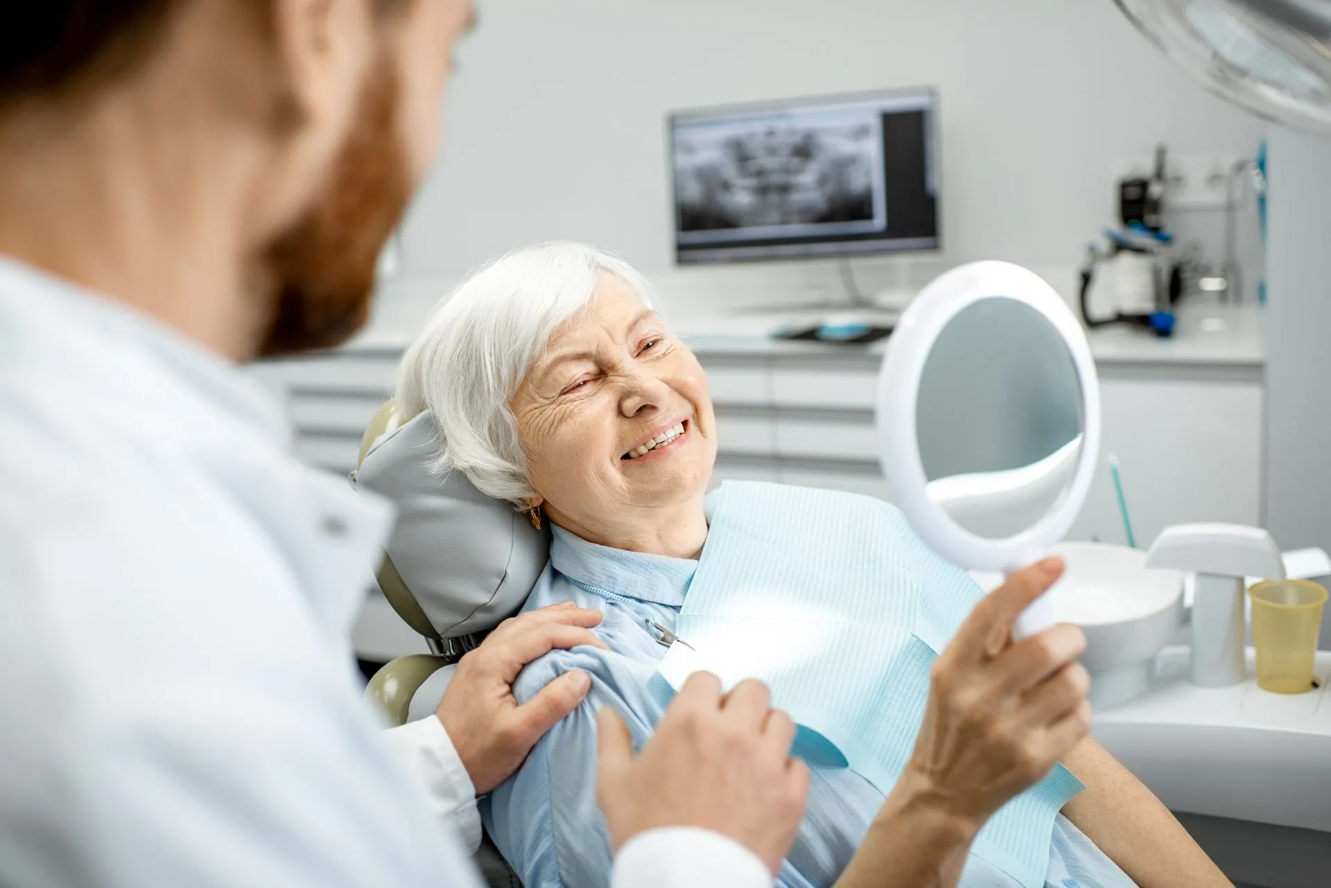 A smiling elderly woman enjoying her beautiful smile, looking in the mirror after denture services.