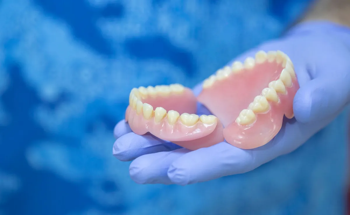 Dentures, pink gums and white teeth, held in a gloved hand.