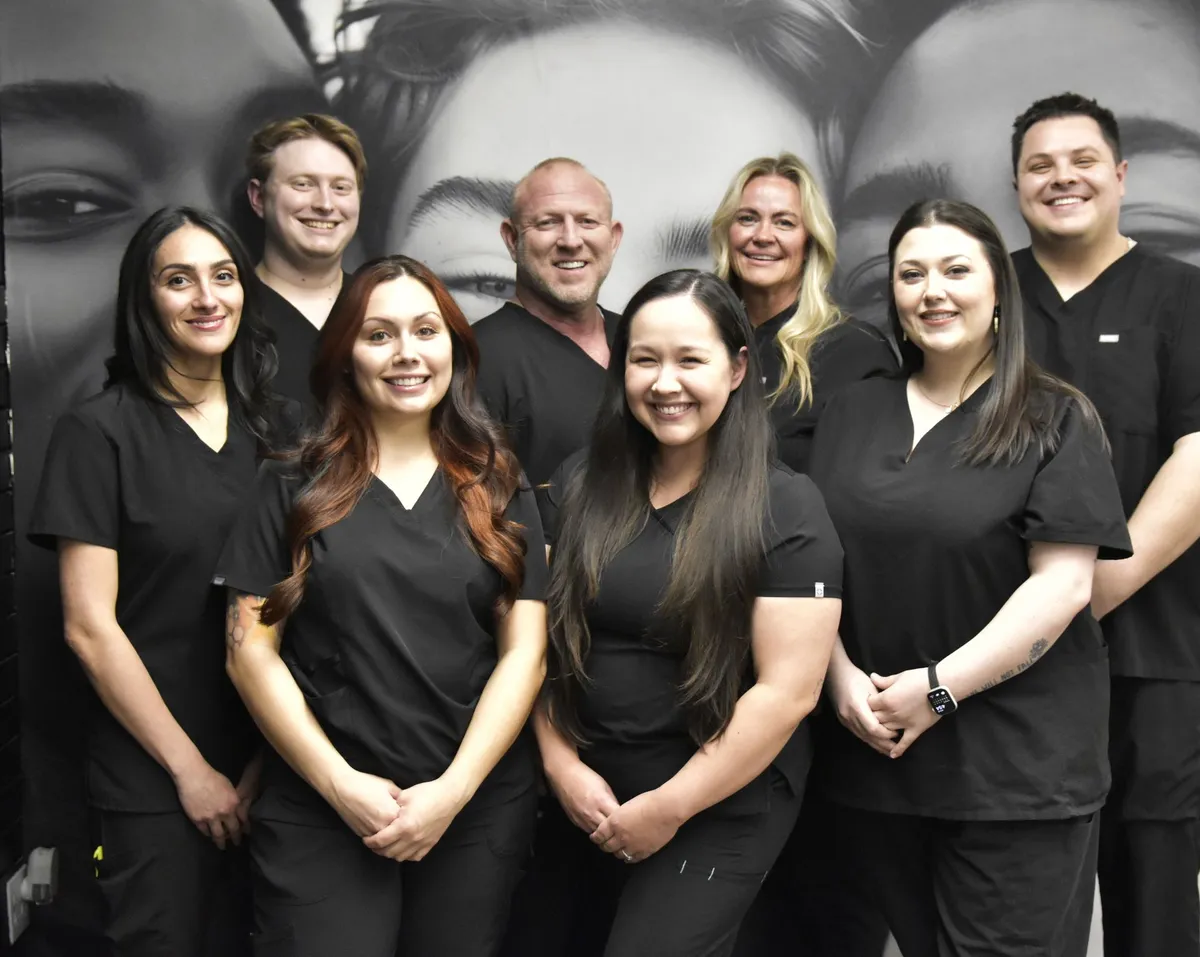 Group of nine people wearing black scrubs pose in front of a green wall and plant.